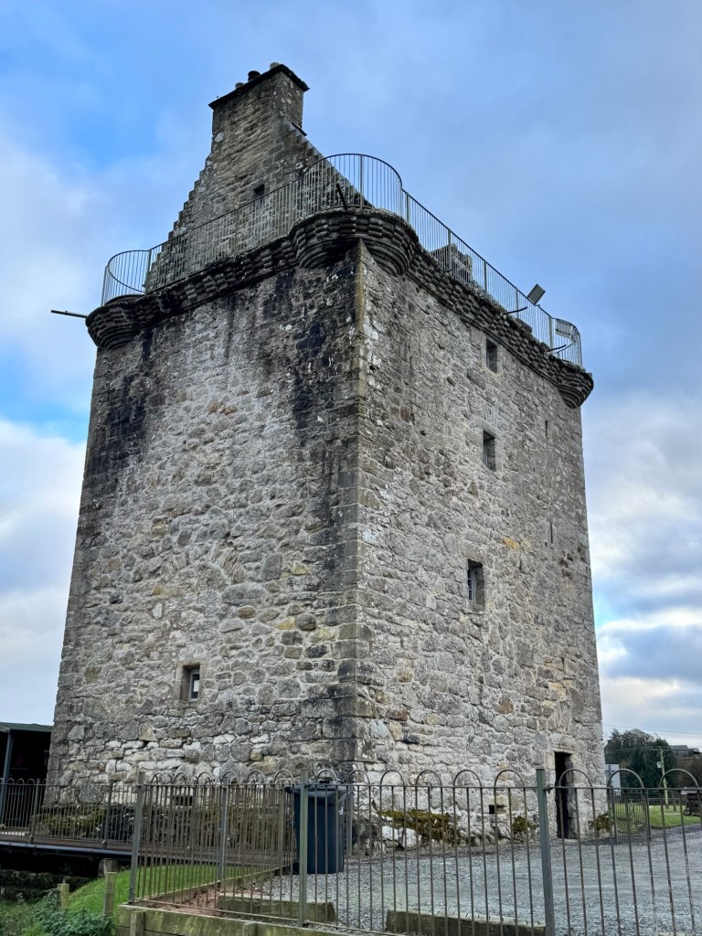 Another well preserved tower in grey stone, viewed from just beyond the car park, Entrance and minimal number of windows visible.