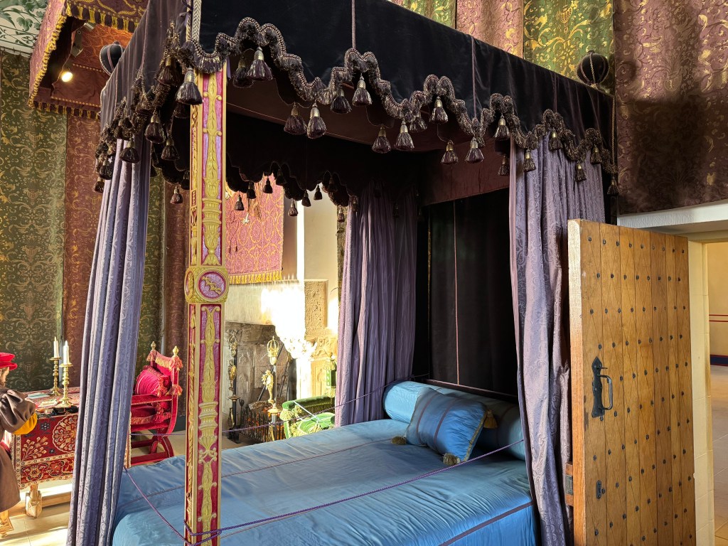 Room, richly decorated with gold, green and claret wall hangings. Four poster bed in centre with opulent looking furnishings in blue and purple.
