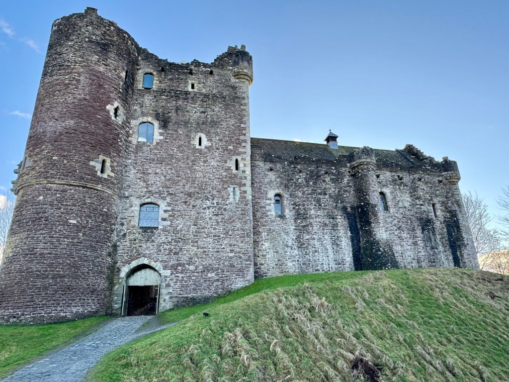 Castle, in grey stone, seen from front, with gatehouse tower to left and great hall to the right. A grassy mound fills the foreground.