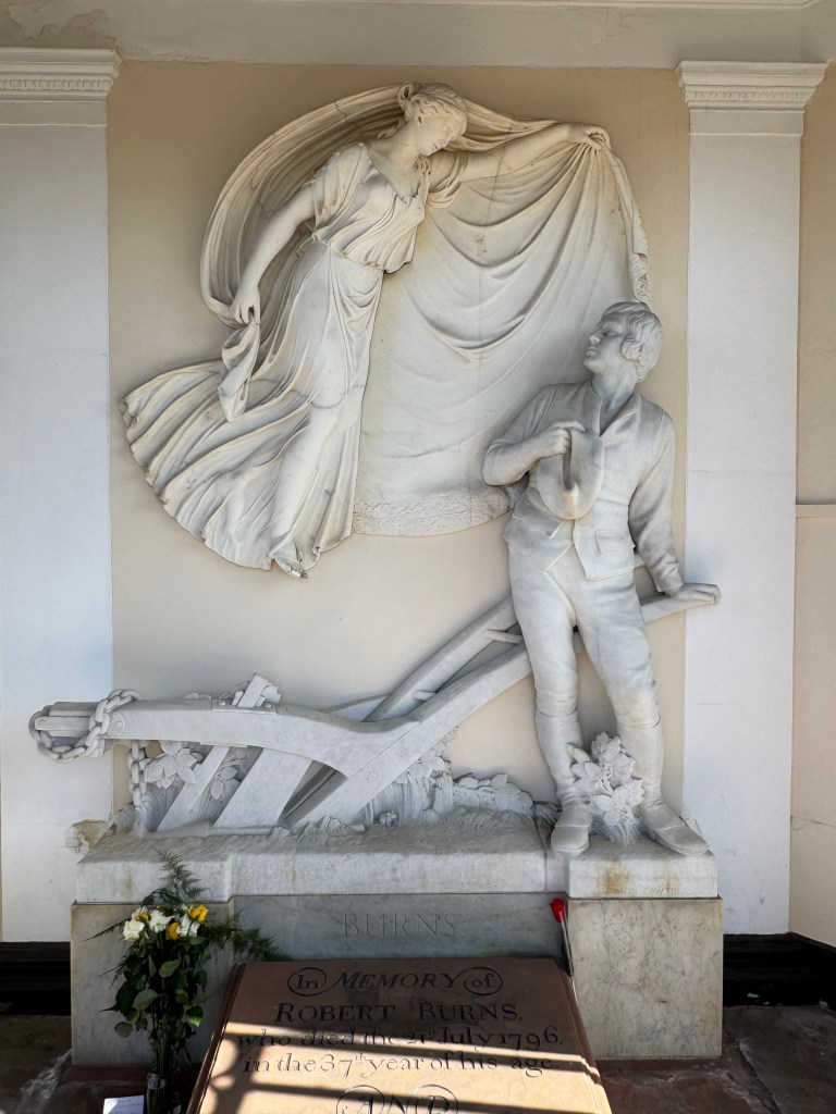 White marble sculpture of Burns in front of ploughshare, with female figure (his muse) hovering above, spreading her cape. Gravestone in the foreground.