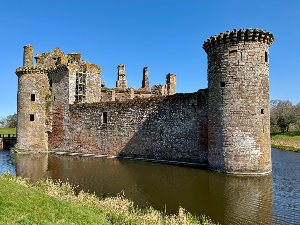 Exterior south west view showing reddish-brown curtain wall, towers and moat. Clear blue sky.