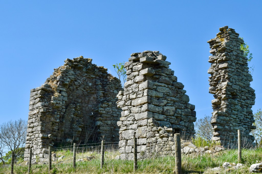 Photo taken below the ruin, looking upwards, showing the severity of the ruin. Wall on south has gone almost entirely. Blue sky.  Ruin is fenced-off.