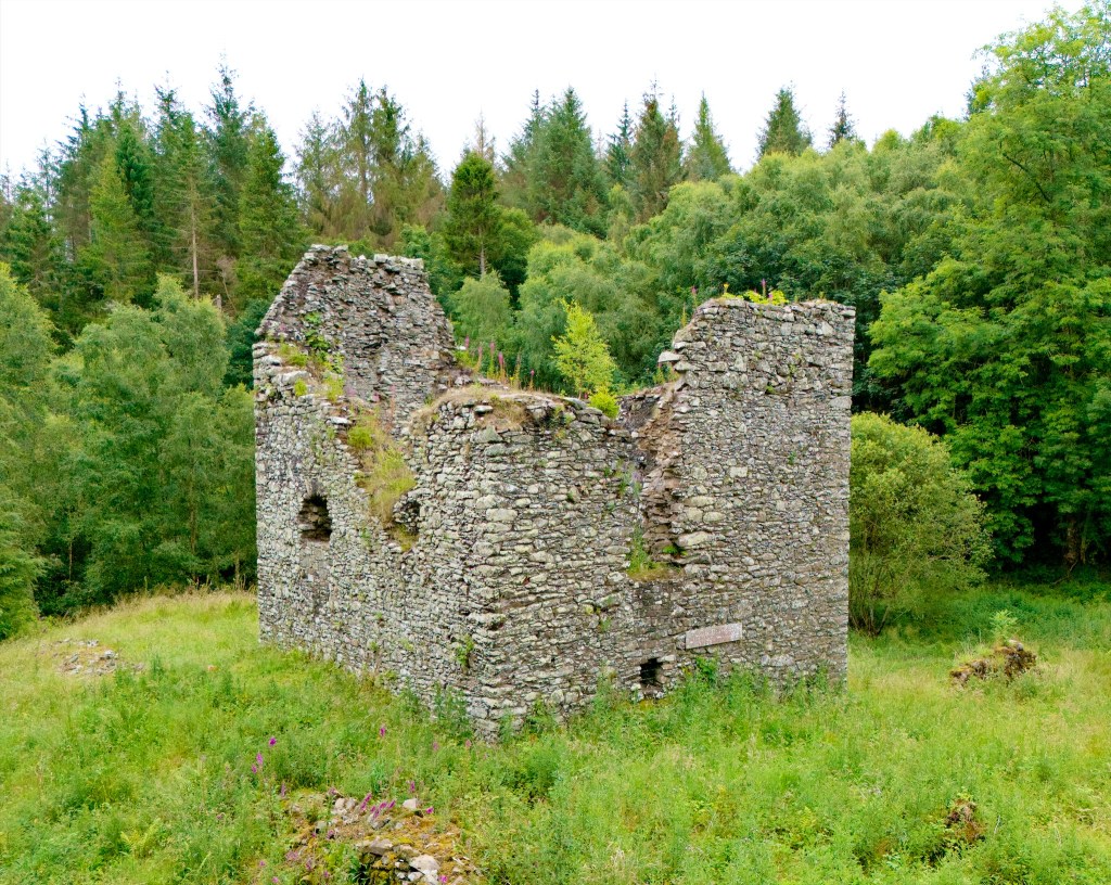 Low level dron shot, showing the ruin against the trees behind. The nearest walls are badly damaged, with lengthy holes running downwards. However, the the visible far wall is almost complete, with the gable end showing.