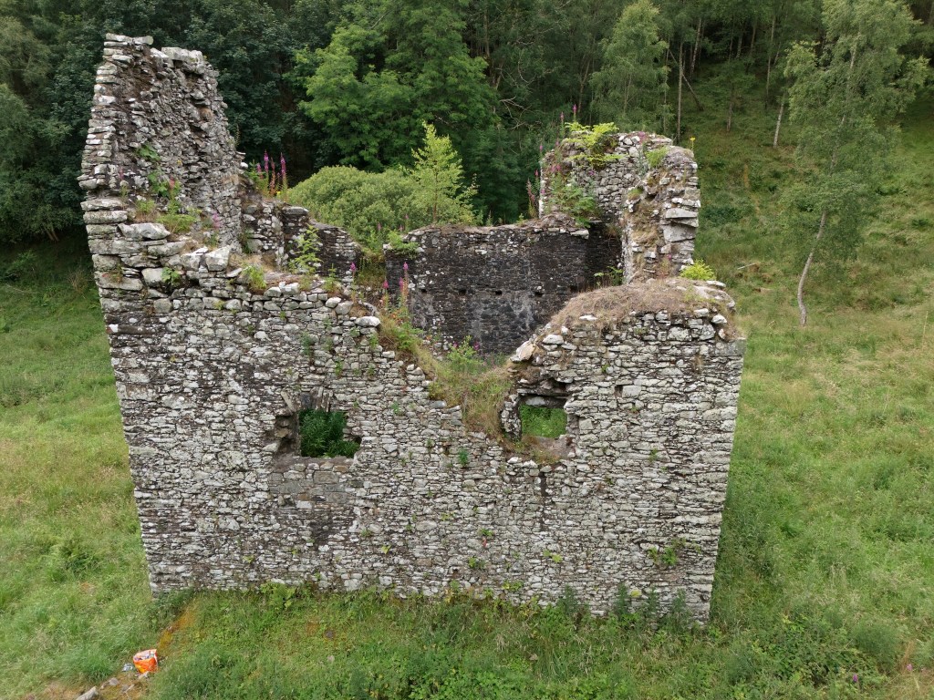 A side-on drone shot, showing the large holes in the facing side wall. The trees are encroaching on the far side.