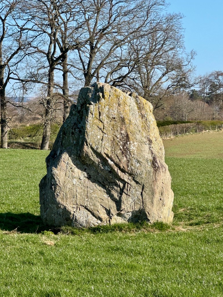 Close-up of a large weathered stone, archlike in shape. Signs of lichen. Trees in background.