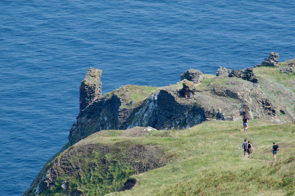 People walking down to the dramatic, cliff-top castle ruin, with deep blue sea in the background. A few framents of castle remain.