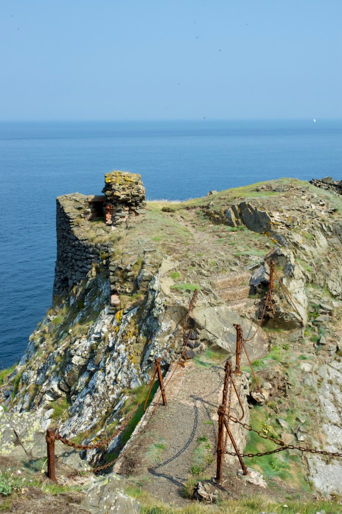 Looking down the gap  that falls away rapidly into the sea. A concrete causeway is visible in the foreground, with rusty chains and fence polls.