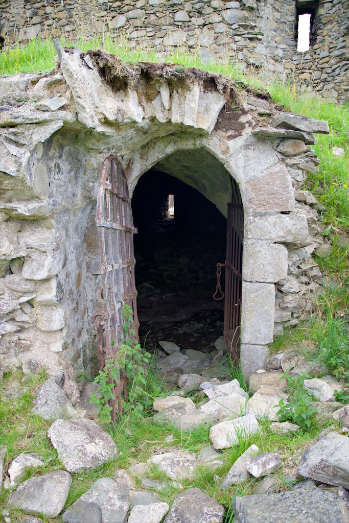 Arched doorway into the basement. Gunloop visible on other side. Lots of rubble around the entrance. Grass growing over the top.