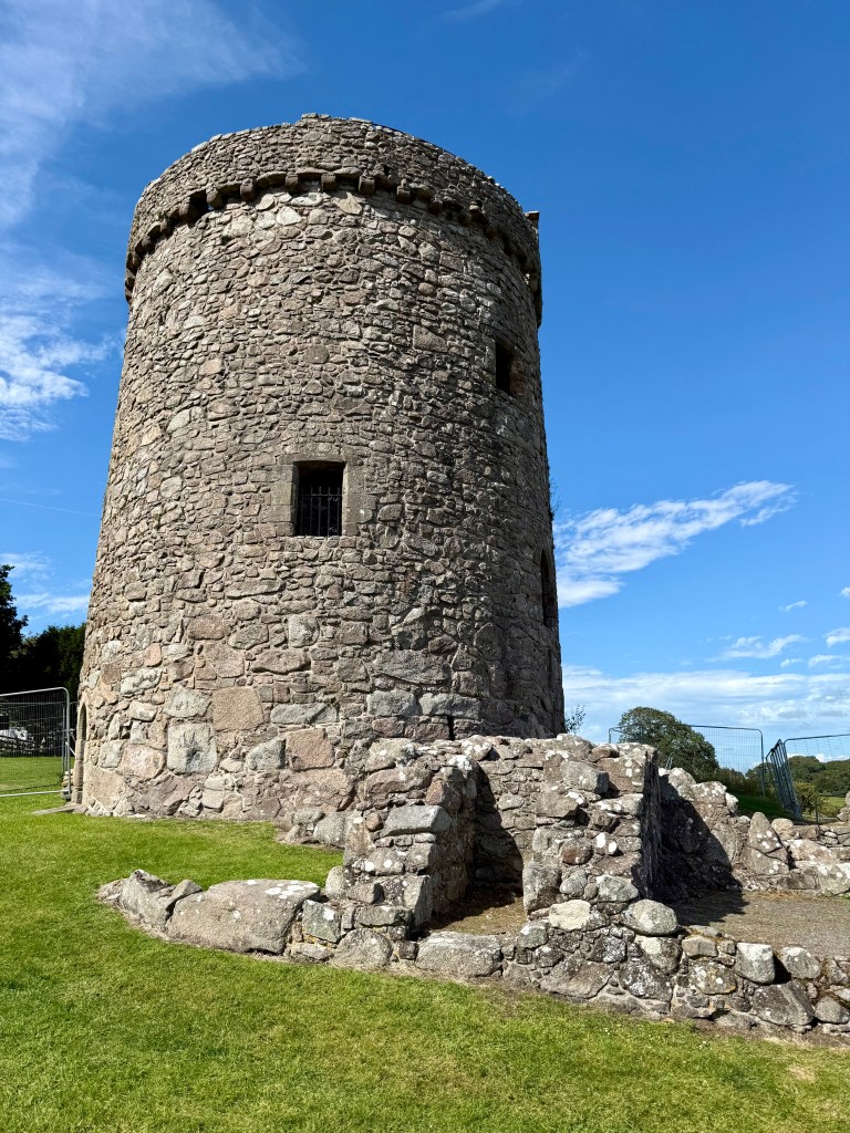 View from south west, with single window in the grey stone half-way up, and remains of outbuildings to the right. All set against a blue sky.