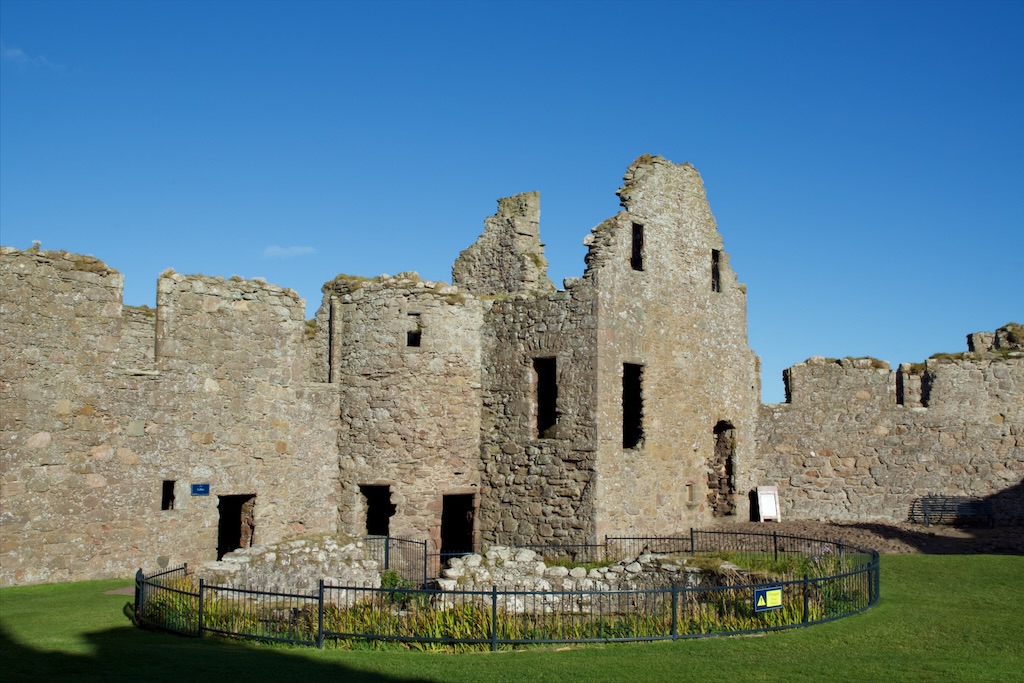 Roofless ruin behind a fenced-off circular area (The Cistern), with some stonework too. Grass in front.