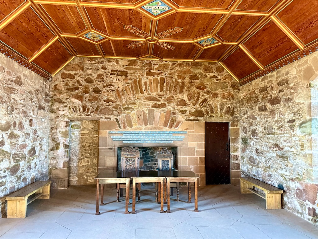 Sone walls and wood panelled ceiling, with gold coloured trimmings. Desk and two chairs in front of fire place. White or greyish tiled floor.