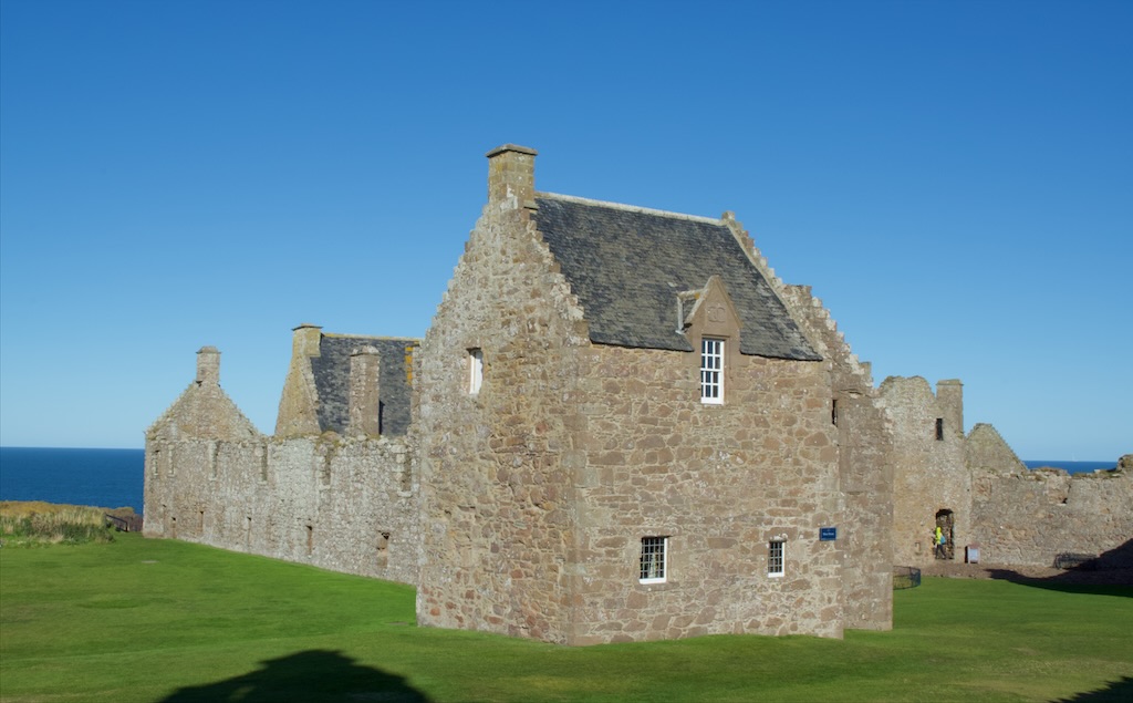 Side view of house, one upstairs window and two downstairs. Wall and building behind, just to roof height, but no actual roof. A setries of windows ar intervals. neatly cut grass surrounds the buildings.
