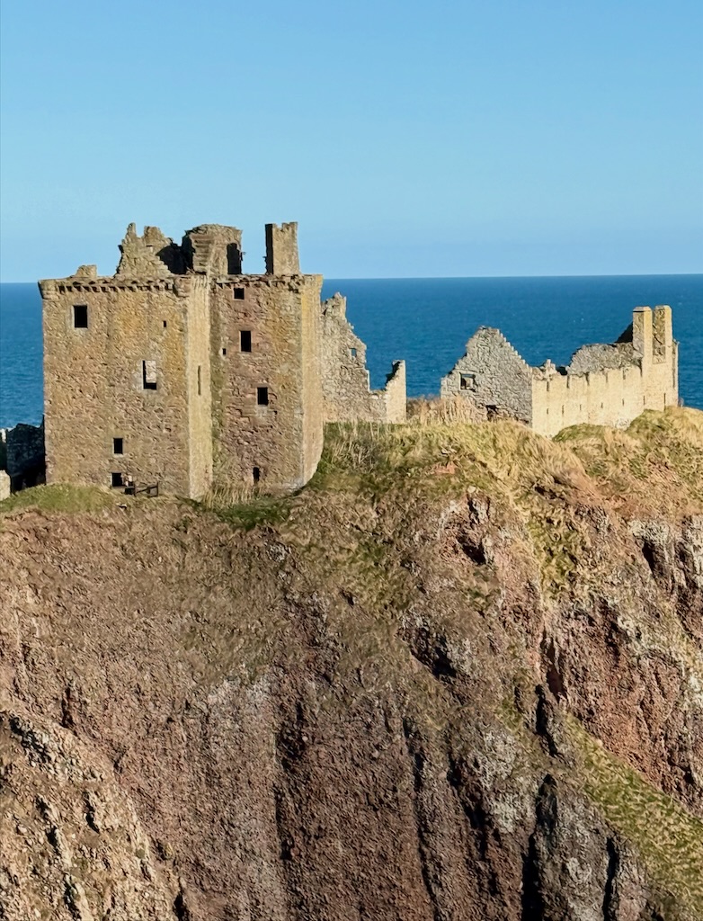 telephoto image from the mainland cliff-top. showing the Keep, right on the edge above the cliffs, falling away to the North Sea below. A clear blue sky behind.