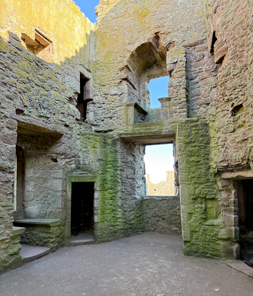 Internal walls, stained with greenish colour. Windows left and centre at camera level. Also winows and openings at level above. No celings or roof. Grey/brown stone dominates structure.