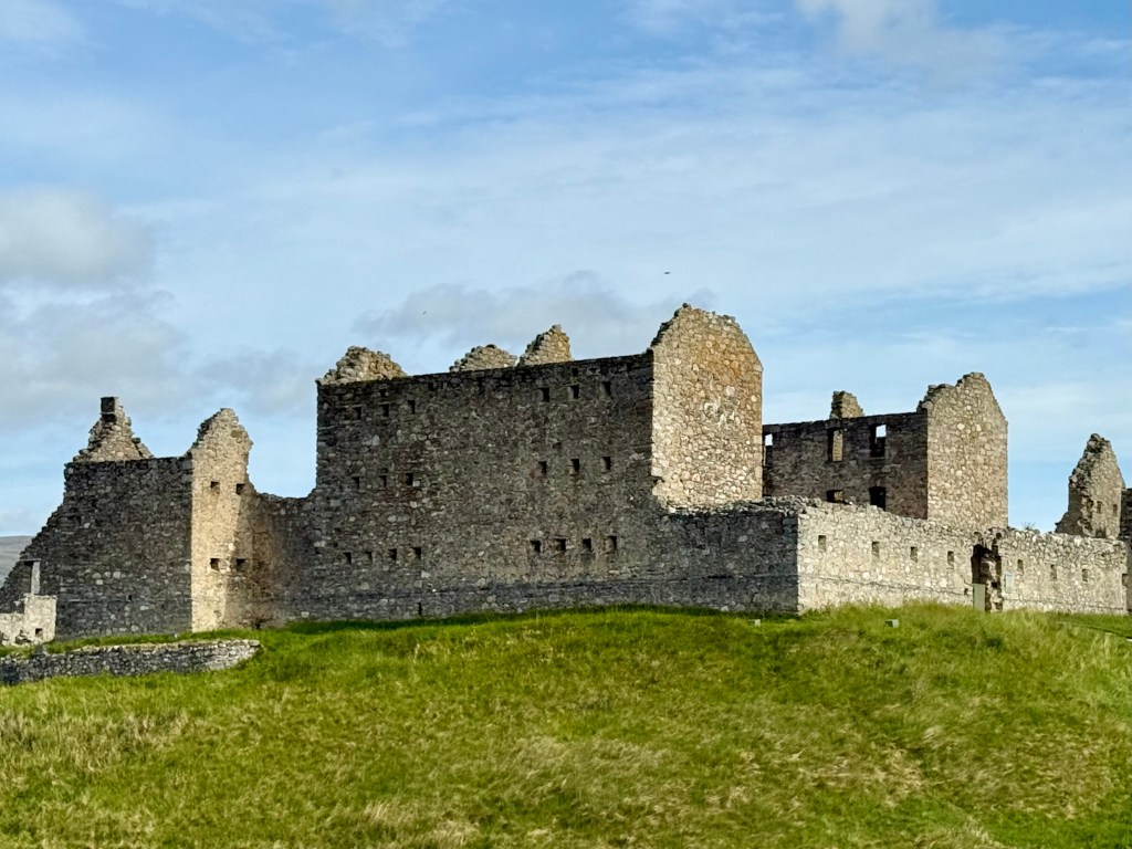 View from roadside, showing outer walls connecting the barrack blocks, with tower towards the left. Hill slopes away below.