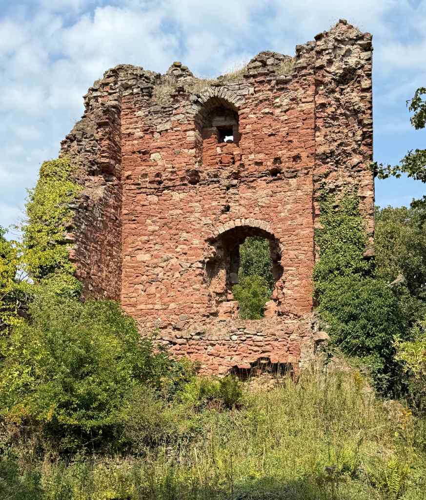 The original entrance at first floor level is clearly visible, with arch over the top. Window above. Overgrown side walls have crumbled. 
