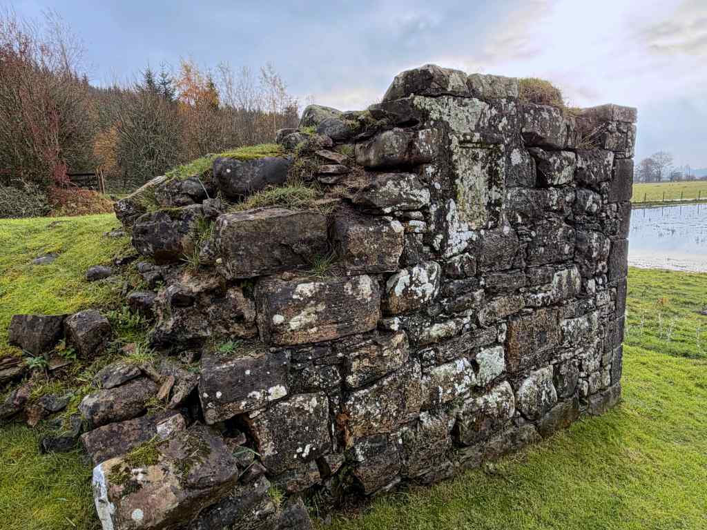Close-up of a larger piece of wall, very weathered, partially restored in corner. Black, grey and brownish coloured stones, overgrown with moss and lichen. Waterlogged field beyond.