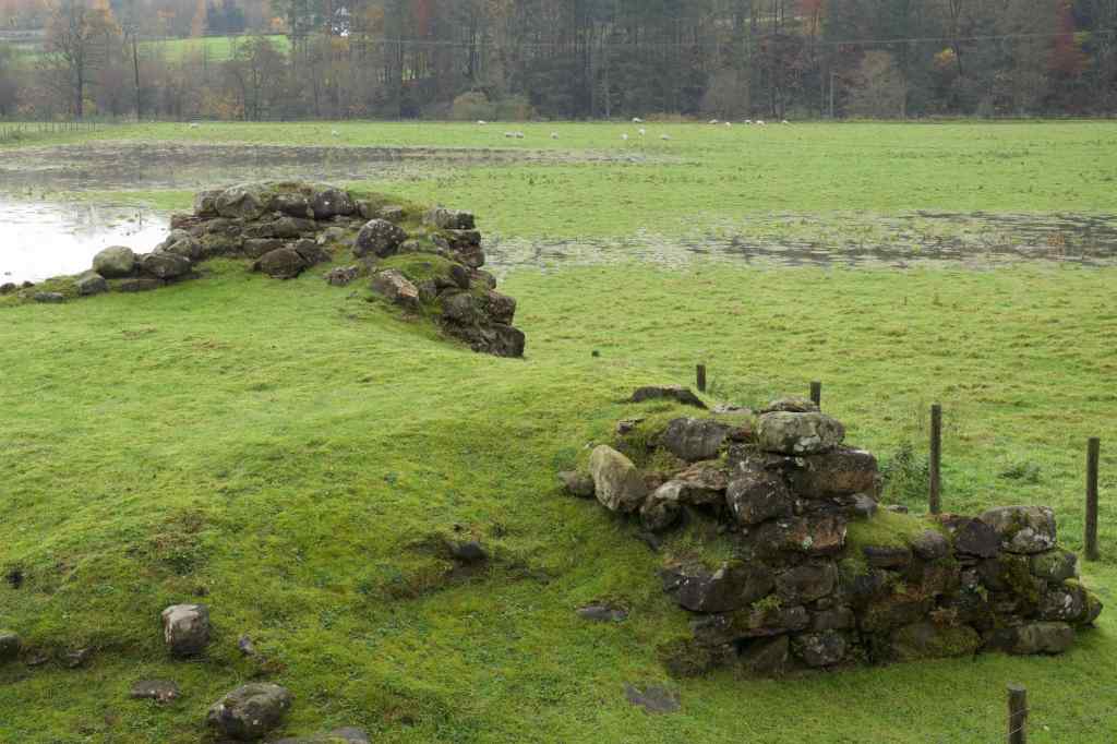 Looking down from railway embankment. Wall remains pushing through grass mound. Waterlogged field behind.