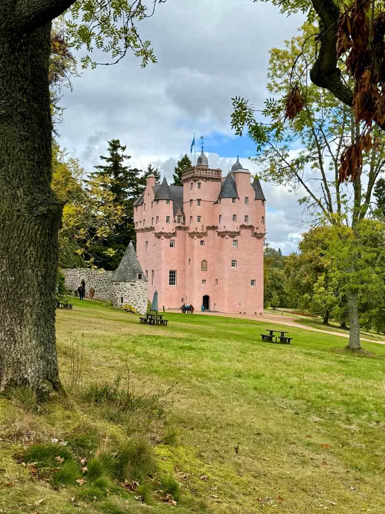 view from the west looking through the trees to the main entrance. Some picnic benches in the foreground. The remaining part of the unharled barmkin wall is also visible.