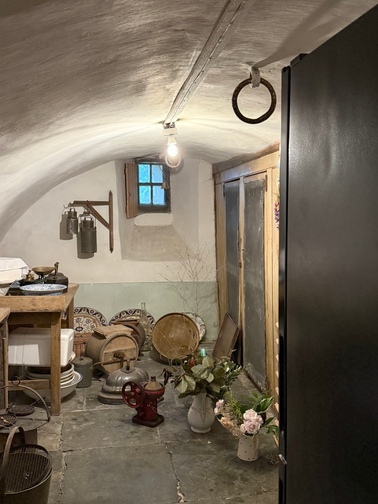Curved roof of valuted basement visible. Painted white. Various country kitchen artefacts on the floor (pots, pans crockery). Very small window in end wall.