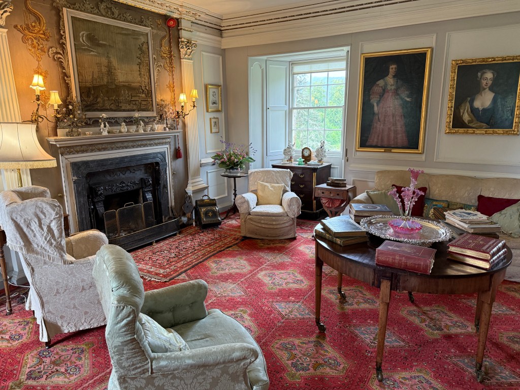 Red carpet with pale green armchairs to left. Small dark wood table, fireplace and portraits on the wall. Window on back wall