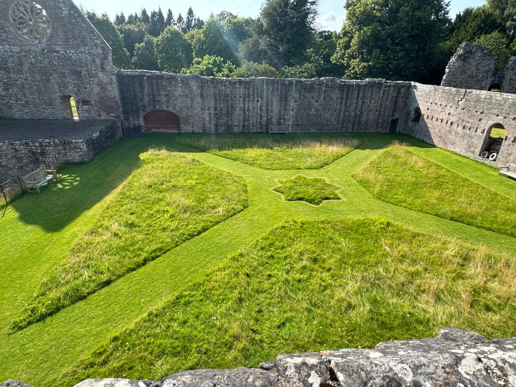 Grassed quadrangle, surrounded by a single-storey wall, except on top left, where it is much taller, being the gable end of a building